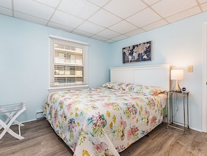 A bedroom with a bed, a window with blinds, a wall-mounted lamp, and a small table with a lamp. - Oceanfront condos with 3 night minimum rentals and linens included! (Ocean City)