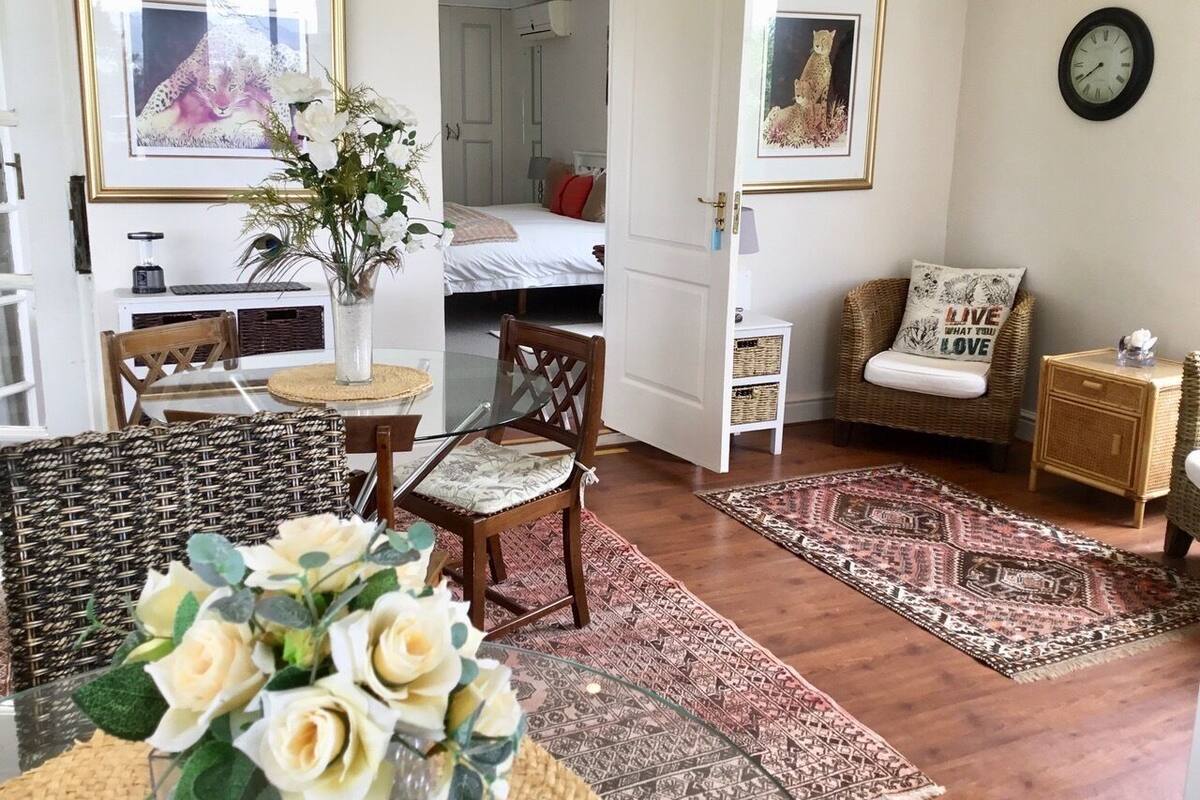 A cozy living room with a dining area, a wicker chair, a glass table, and a vase of flowers.