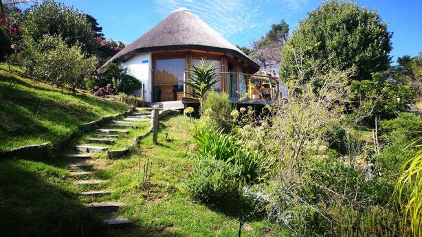 A thatched-roof house with a wooden deck, surrounded by lush greenery and a clear blue sky. - Ocean Golf Guest House (Cape Town)