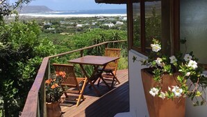 A wooden deck with a table and chairs, overlooking a coastal landscape with a view of the sea and a mountain in the distance. - Ocean Golf Guest House (Cape Town)