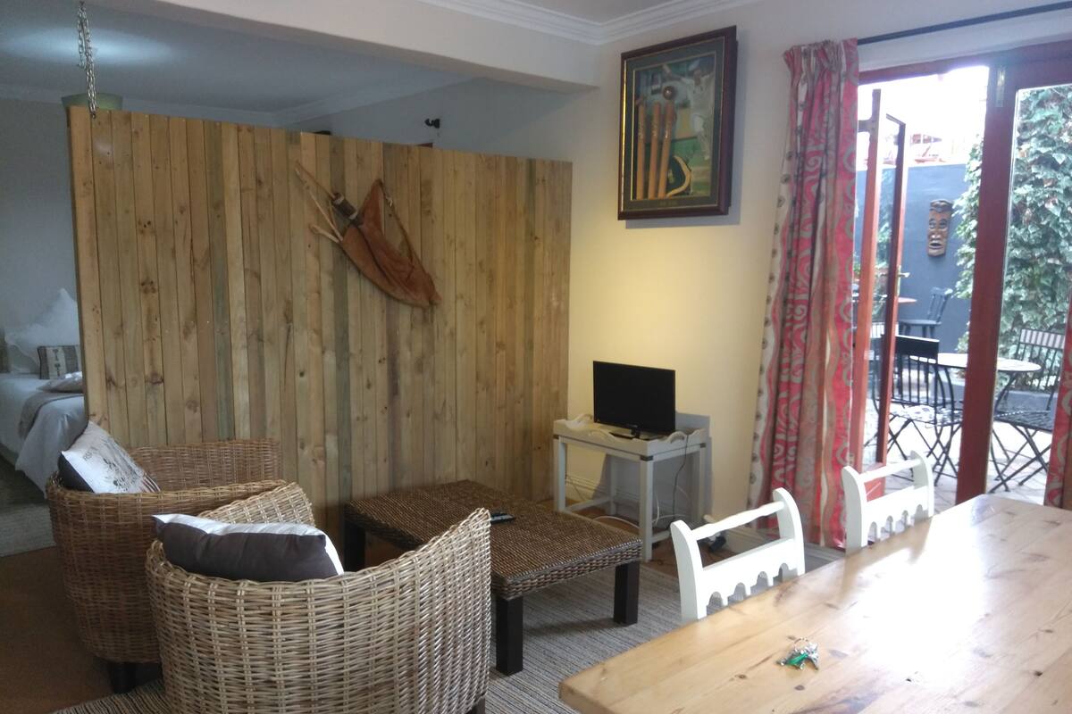 A cozy living room with wicker furniture, a wooden wall, a TV on a small table, and a balcony with chairs.
