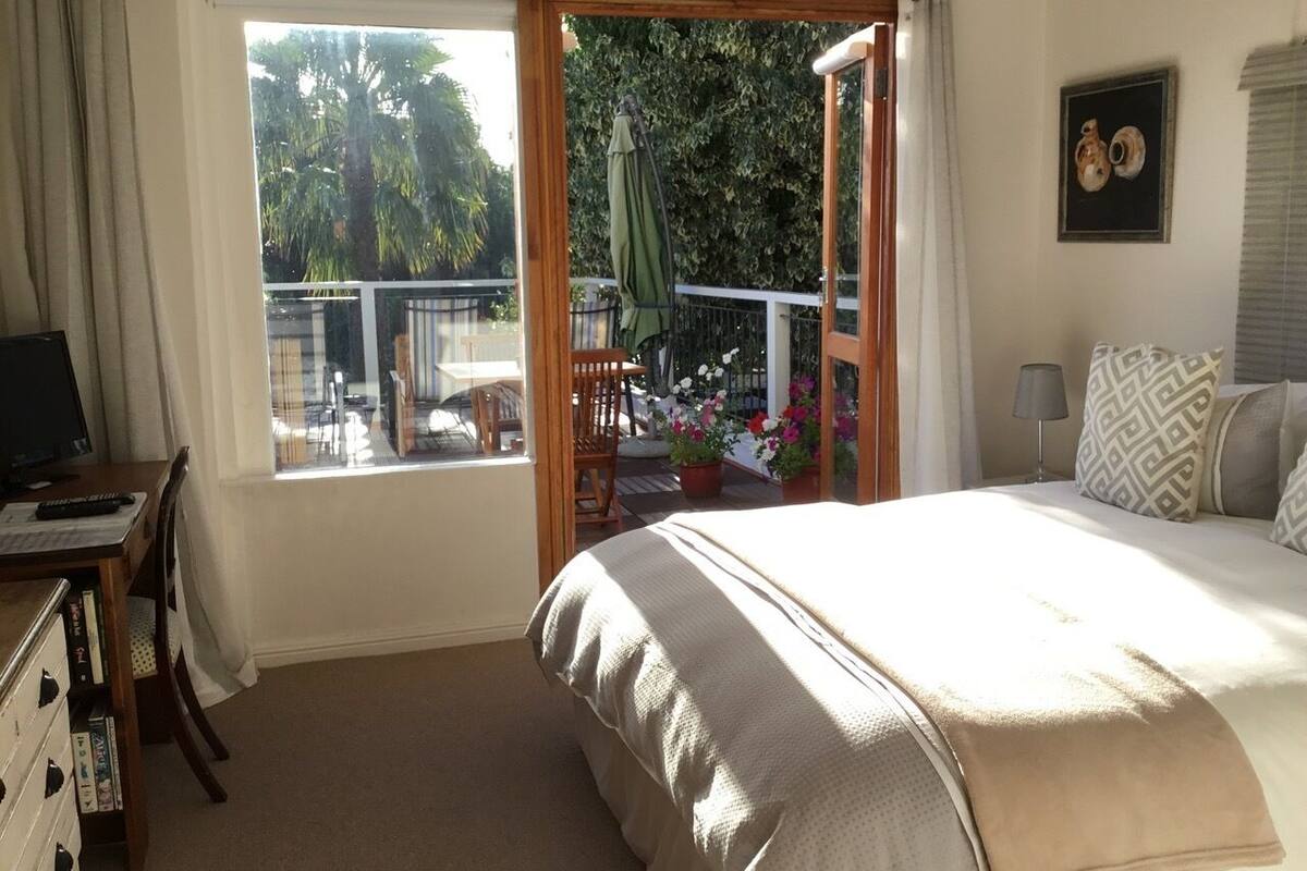 A bedroom with a large bed, a desk with a computer, and a view of a balcony with plants and a chair.
