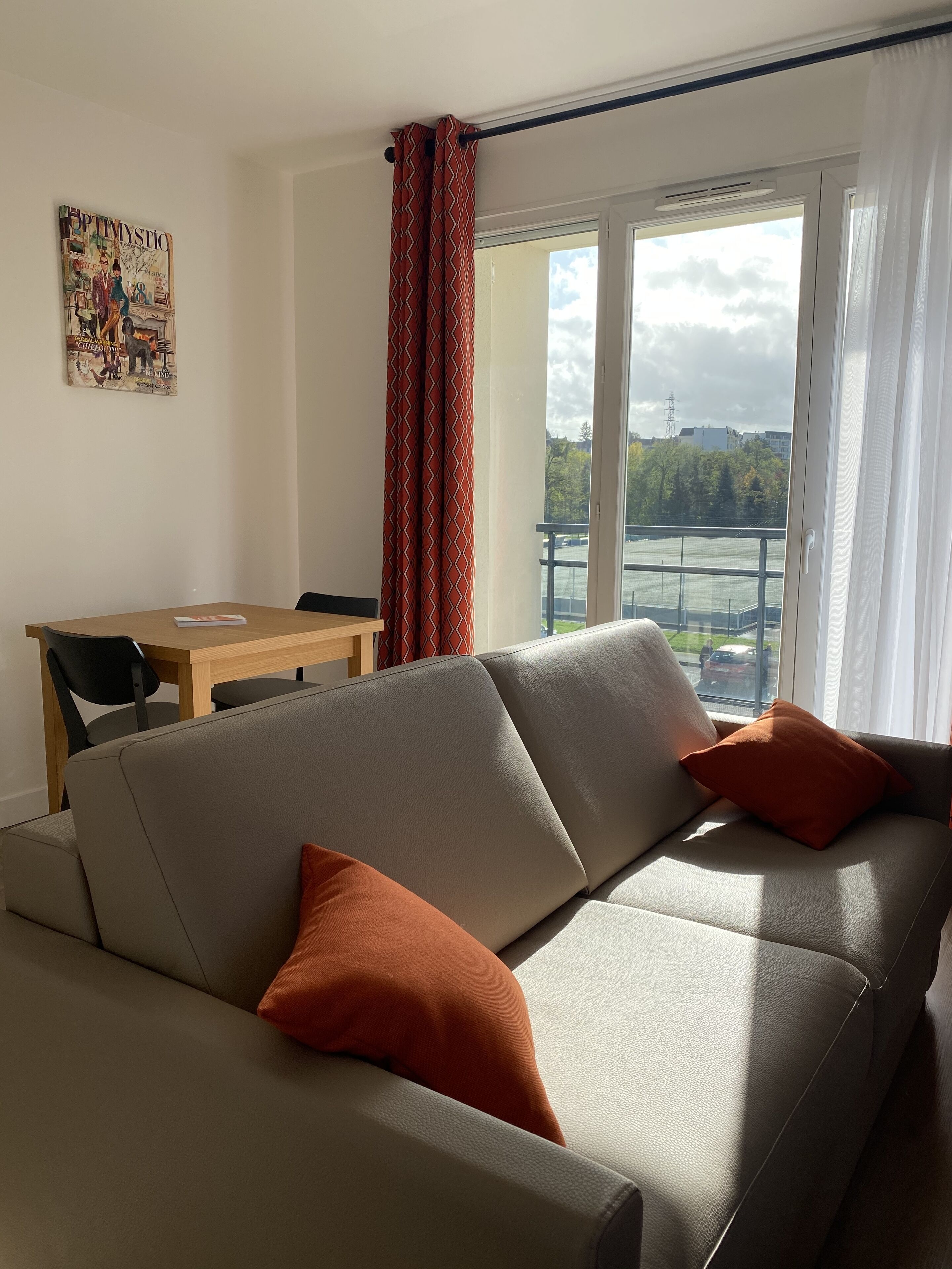 A living room with a sofa, two orange cushions, a wooden table with chairs, and a window with curtains.