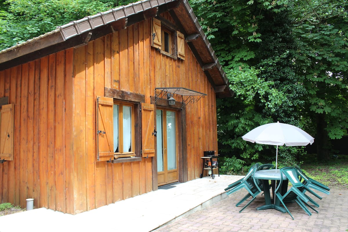 A wooden cabin with a brick pathway leading to a table and chairs under a white umbrella.