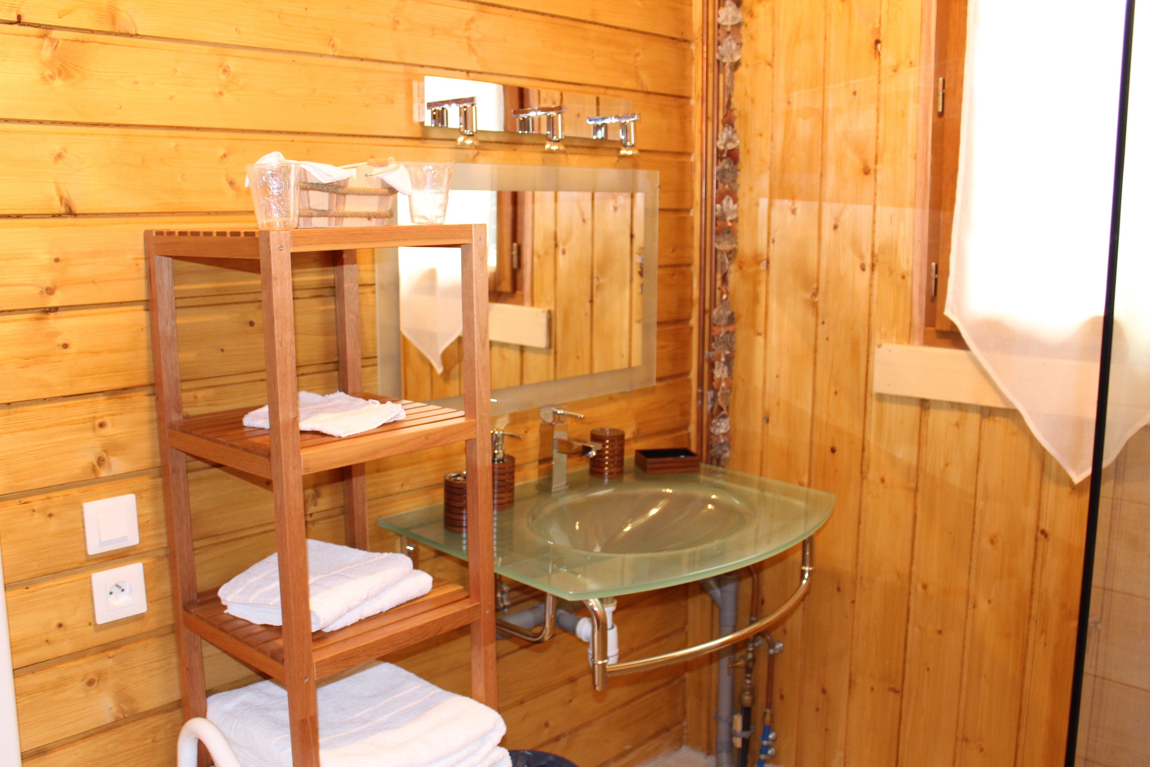 Une salle de bain rustique, avec une cabine en bois, un lavabo en verre, des étagères en bois où sont posées des serviettes et un miroir au-dessus du lavabo.