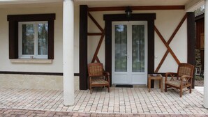A covered patio area with two wicker chairs and a small table, a door with glass panels, and a window with a cup on the ledge. - Sawan (Jâlons)