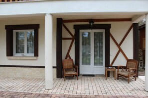 A covered patio area with two wicker chairs and a small table, a door with glass panels, and a window with a cup on the ledge. - Sawan (Jâlons)