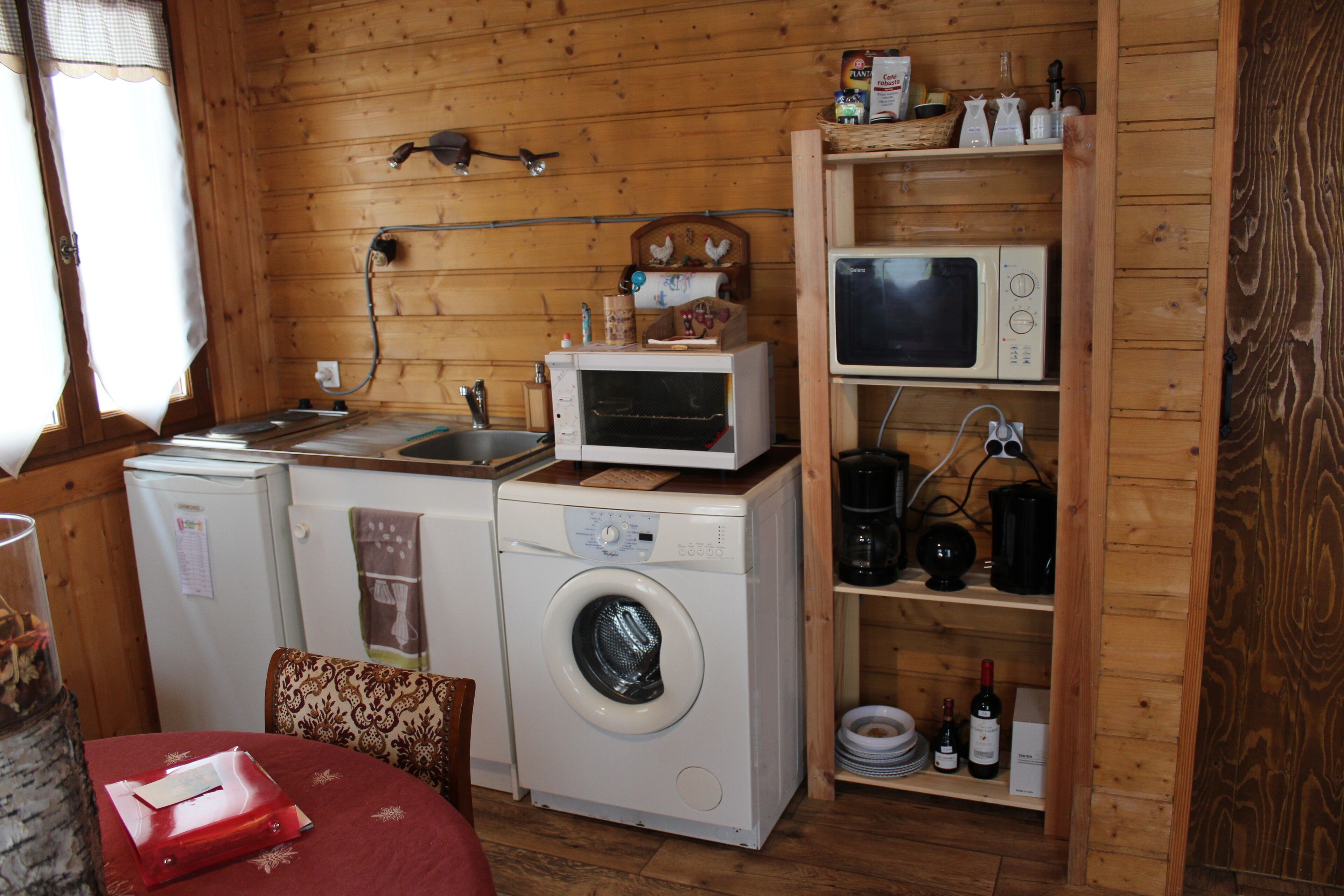 A compact kitchen with a washing machine, microwave, and a shelf with kitchenware and wine bottles.