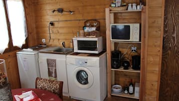 A compact kitchen with a washing machine, microwave, and a shelf with kitchenware and wine bottles.