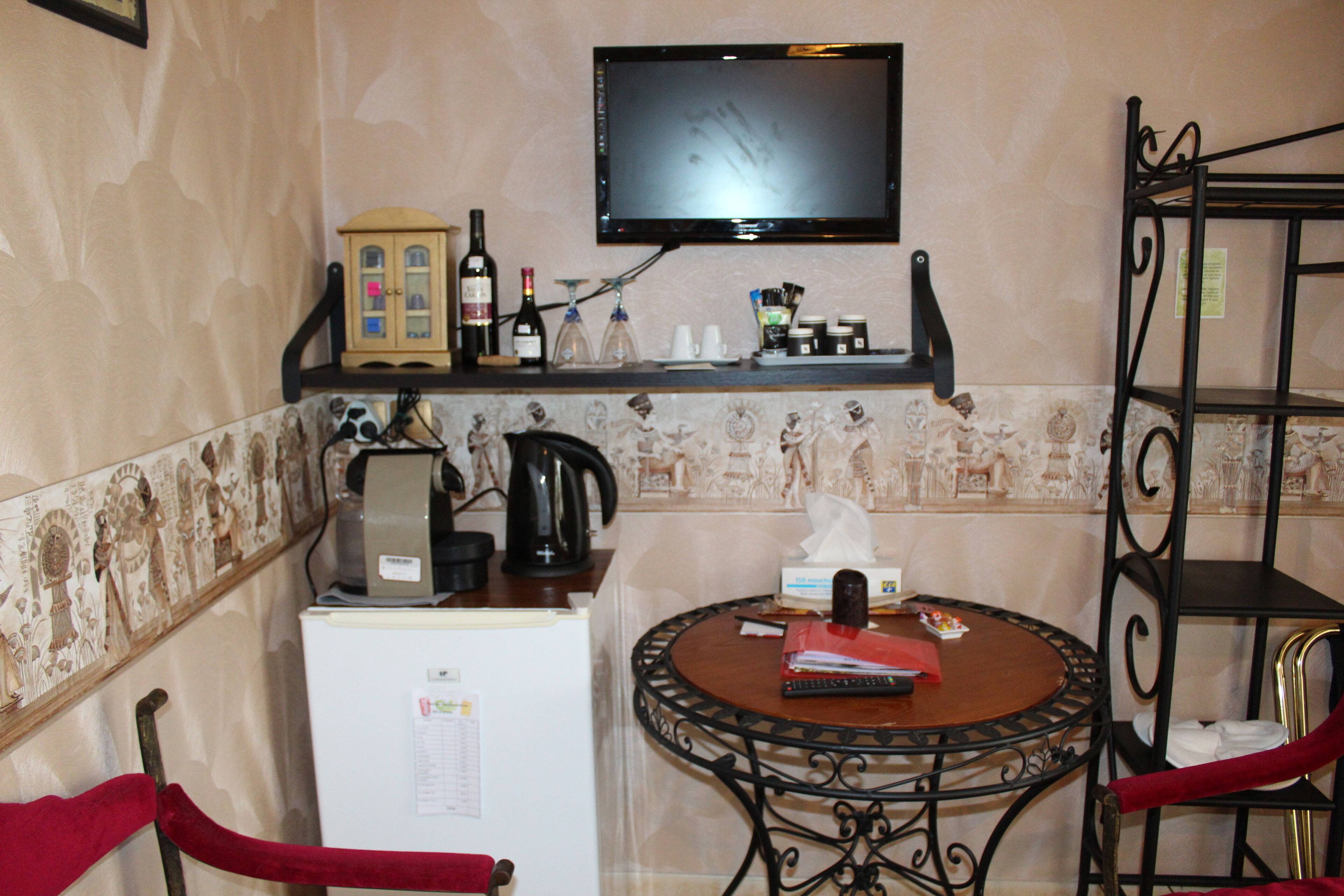 A small kitchen with a refrigerator, a coffee maker, a wine bottle, and a television mounted on the wall.