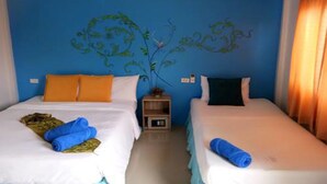 A hotel room with two beds, each with white bedding and blue towels. There is a small nightstand with a lamp and a decorative plant. The walls are painted blue, and there is a window with curtains. - Lanta at Lanta Place (Ko Lanta)