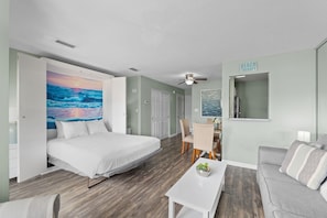 A bedroom with a bed, a desk, a chair, a sofa, a ceiling fan, and a wall with a beach therapy sign.