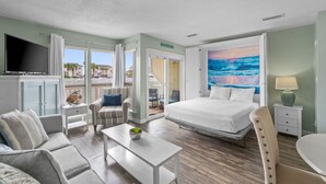 Una camera d'albergo con un letto, una televisione, un divano, una poltrona, un tavolino e un quadro raffigurante l'oceano.