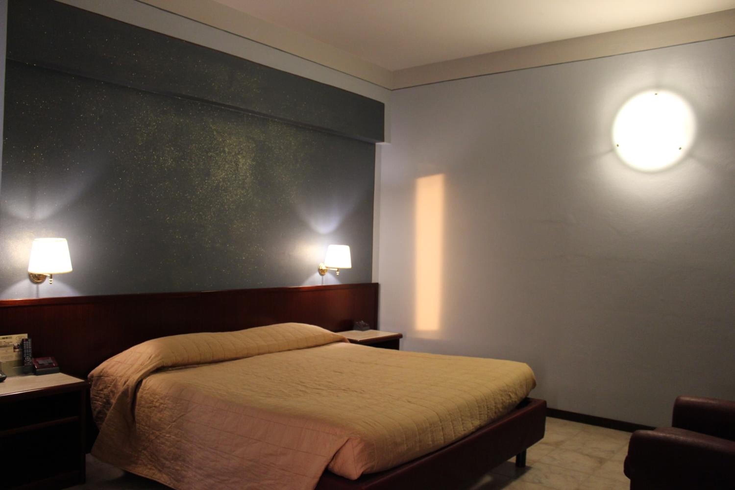 Habitación de hotel con una cama grande, dos lámparas de noche, una mesita de noche y una lámpara de pared redonda.