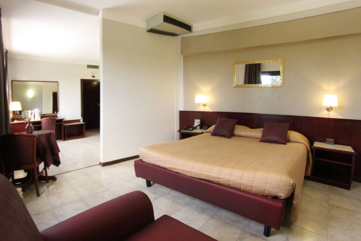 A hotel room with a large bed, a desk, a chair, a bedside table, and a mirror on the wall.