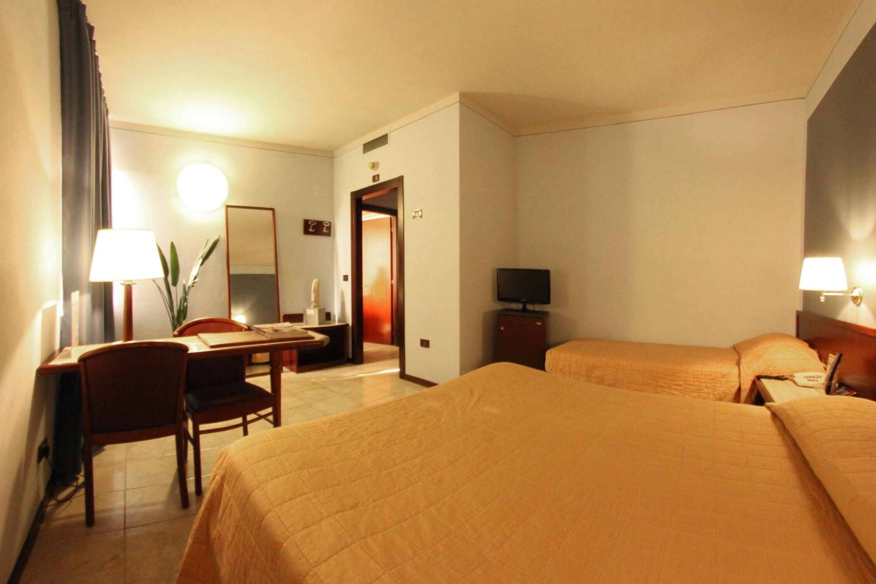 A hotel room with two beds, a desk, a chair, a TV, and a lamp.