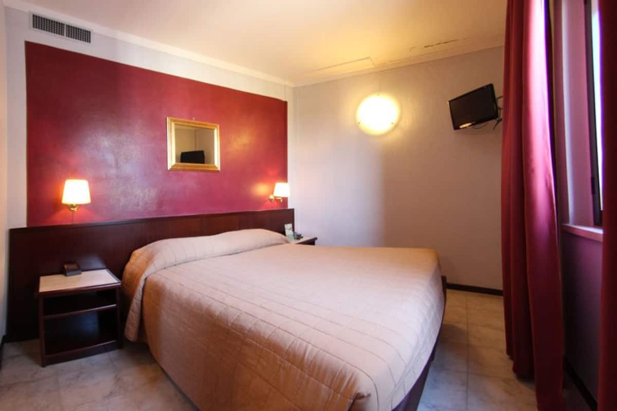 A hotel room with a bed, two bedside lamps, a nightstand, a mirror, a television, and a red curtain.