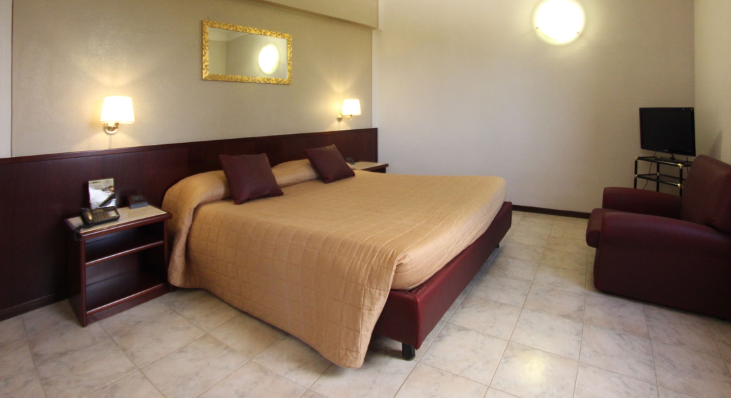 A hotel room with a large bed, bedside tables, a TV, and a red armchair.