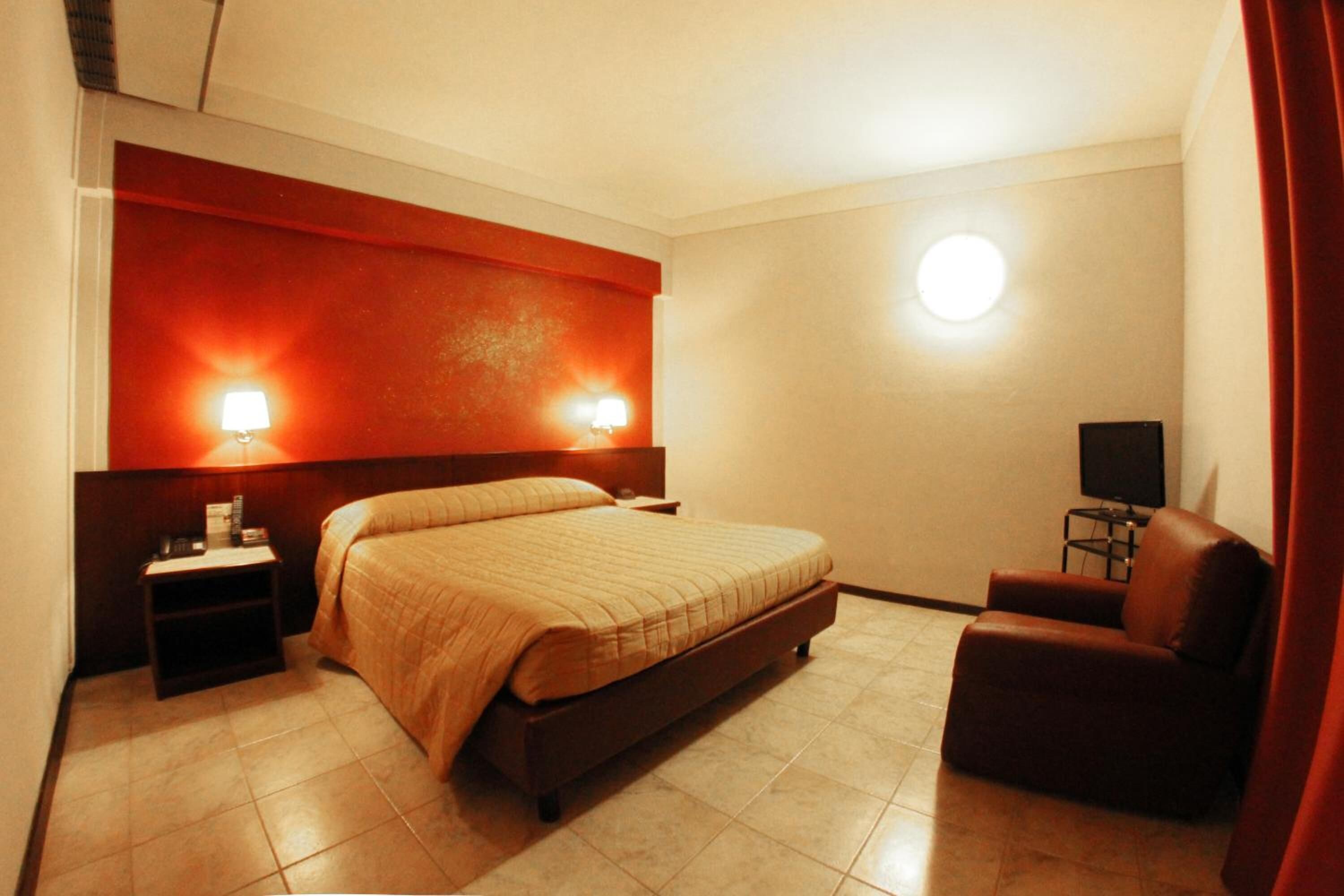 Ein Hotelzimmer mit einem Bett, einem Nachttisch, einem Sessel, einem Fernseher und einer Wandleuchte.