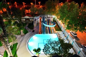 Una piscina al aire libre con tobogán, rodeada de árboles y un camino pavimentado.