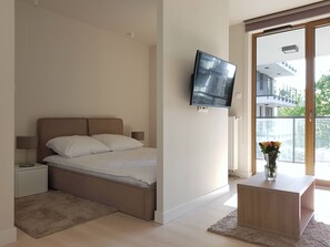 Un dormitorio moderno con una cama, un televisor montado en la pared, una mesita de noche, una pequeña mesa con un jarrón de flores y vistas a un balcón y un edificio.