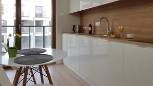 A modern kitchen with a white countertop, a sink, and a wooden backsplash. There is a dining area with a white table and chairs, and a view of the outside balcony. - Chopin Apartments Union (Warsaw)