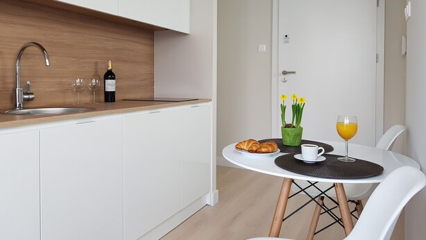 A modern kitchen with white cabinets, a wooden countertop, and a sink. There is a dining area with a white table, two chairs, a plate of croissants, a glass of orange juice, and a vase with yellow flowers. - Chopin Apartments Union (Warsaw)