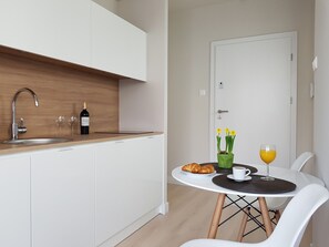 Eine moderne Küche mit weißen Schränken, einer Holzauskładung und einem Spülbecken. Es gibt eine Essecke mit einem weißen Tisch, zwei Stühlen, einer Croissantplatte, einem Glas Orangensaft und einer Vase mit gelben Blumen.