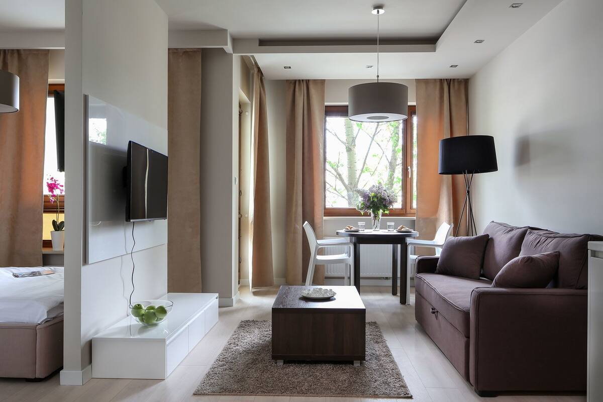 A modern hotel room with a flat-screen TV, a sofa, a dining table with chairs, a coffee table, and a lamp.