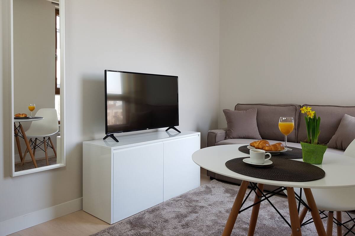 A modern living room with a flat-screen TV on a white cabinet, a round dining table with a placemat, a sofa, and a coffee table with breakfast items.