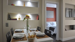 Un comedor con una mesa dispuesta para cuatro, incluyendo platos, vasos y una cesta de pan. Hay libros y objetos decorativos en estantes sobre la mesa.