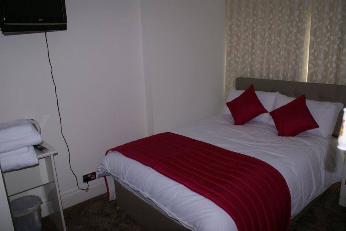 Ein ordentlich bezogenes Bett mit einer roten und weißen Tagesdecke, ein weißer Nachttisch mit Handtuch und Mülleimer sowie ein an der Wand montierter Fernseher.