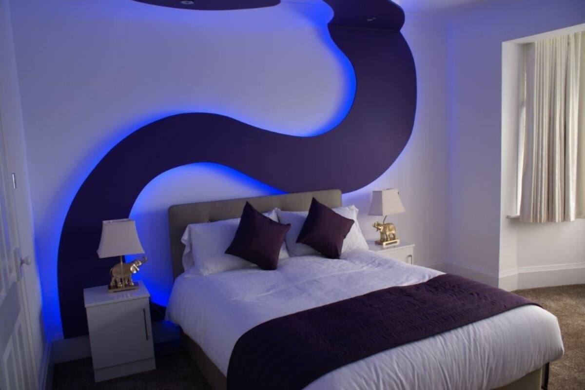 Ein modernes Schlafzimmer mit einer gewölbten Wandgestaltung, einem großen Bett mit weißer Bettwäsche und violetten Akzenten, zwei Nachttischen mit Lampen und einem Fenster mit Vorhängen.