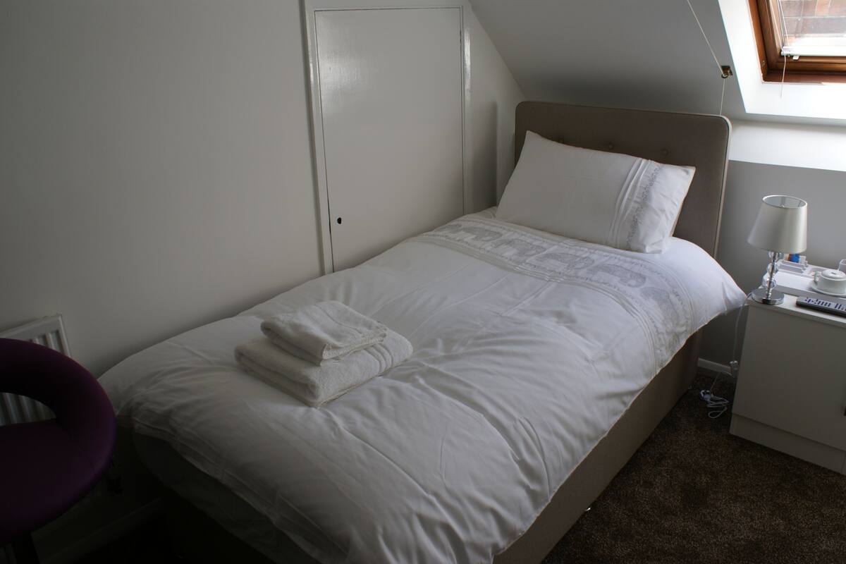 Ein ordentlich bezogenes Bett mit weißen Leinen und einem gefalteten Handtuch, ein Nachttisch mit Lampe und ein Fenster mit Blick ins Freie.