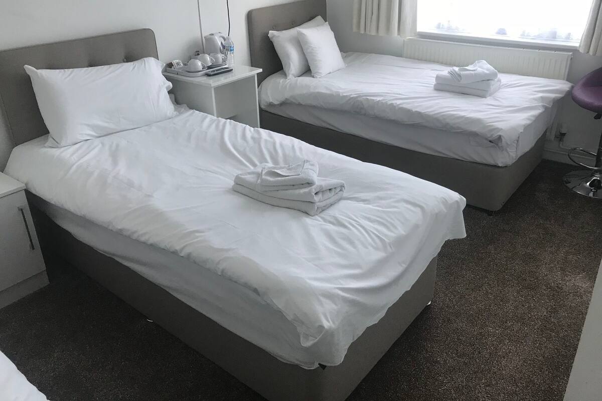 Ein Hotelzimmer mit zwei Betten, jeweils mit weißer Bettwäsche und Kissen, einem Nachttisch mit Wasserkocher, Fernbedienung und einer Wasserflasche.