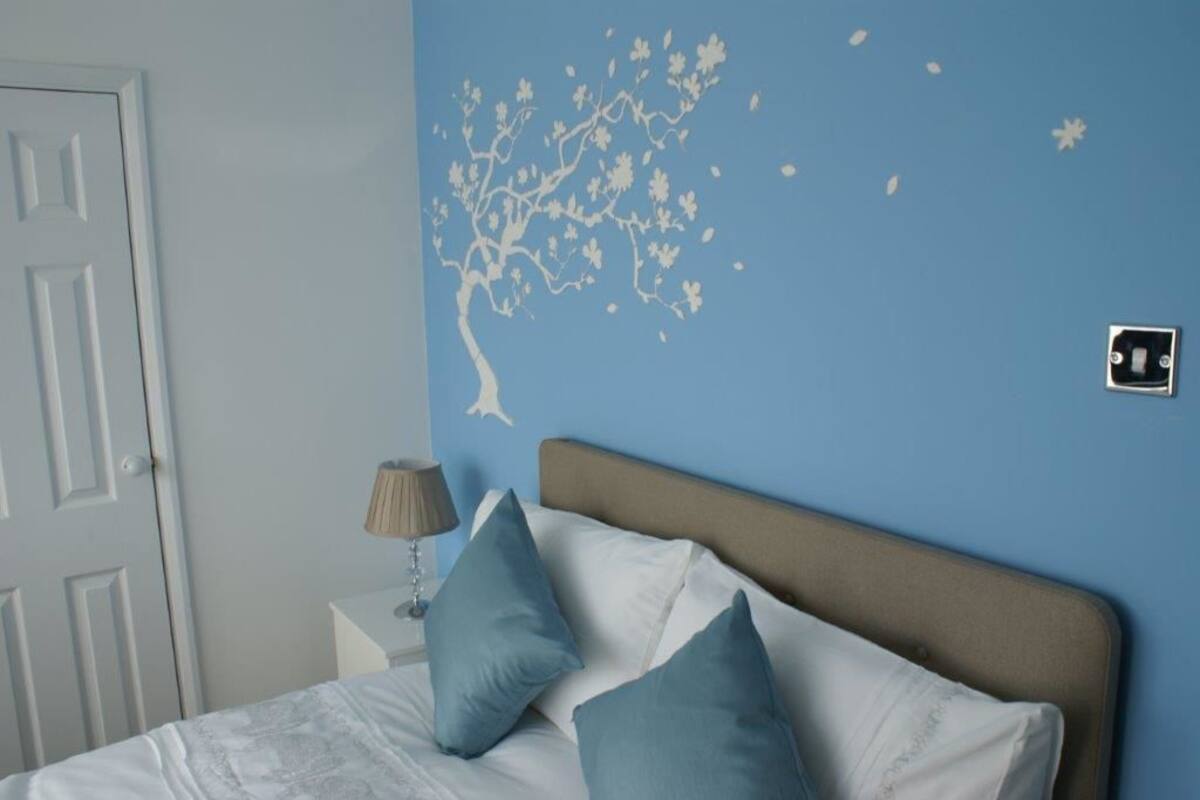 Ein Schlafzimmer mit einem Bett, Kissen, einem Nachttisch, einer Lampe und einer Wand mit einem Wandtattoo von einem Baum.