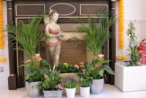 Una estàtua d'una dona amb vestimentes tradicionals, envoltada de plantes i flors en testos davant d'una porta de fusta.