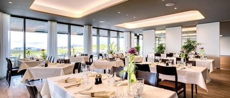 Un restaurant doté de nombreuses tables dressées pour le service, de grandes fenêtres offrant une vue sur l’extérieur et d’un décor intérieur moderne.