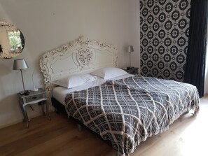 A bedroom with a white ornate headboard, a patterned bedspread, a bedside table with a lamp, and a patterned curtain. - Bed and Breakfast Mooi Achel (Hamont-Achel)