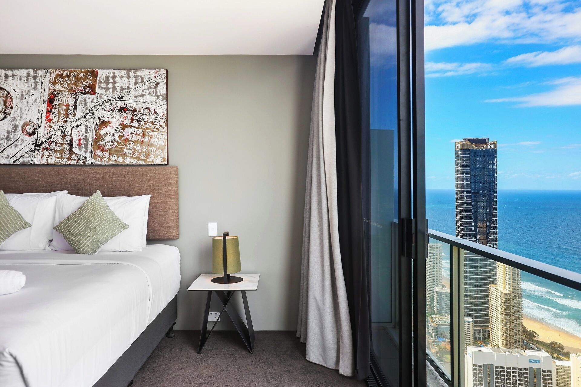 A hotel room with a bed, a small table with a lamp, a large window with a view of the ocean and a tall building.
