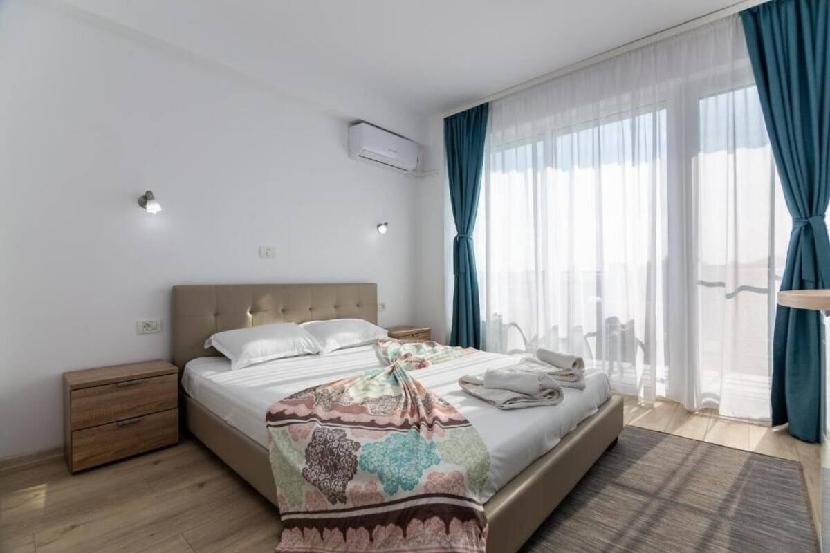 Un dormitorio moderno con una cama grande, una mesita de noche, un equipo de aire acondicionado y vistas al exterior a través de cortinas translúcidas.