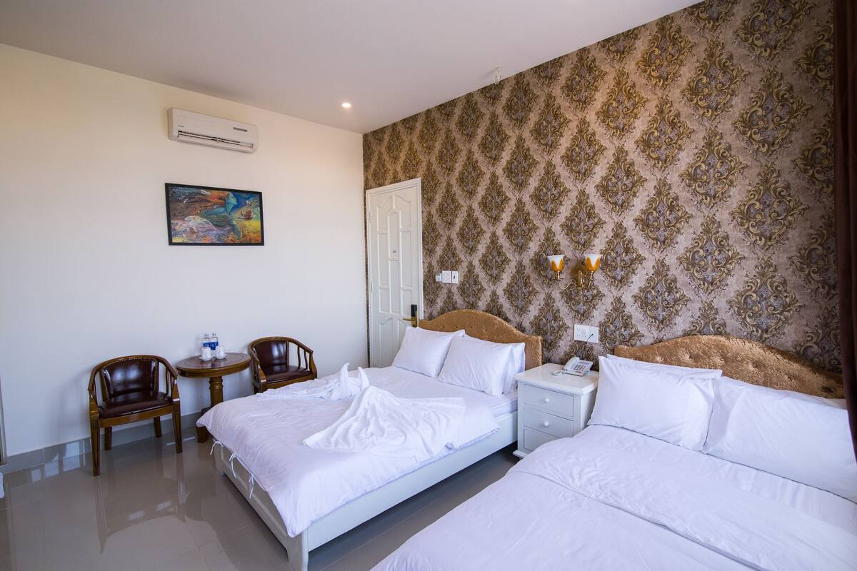 A hotel room with two beds, a wooden chair, a small table, an air conditioner, a painting, and a patterned wallpaper.