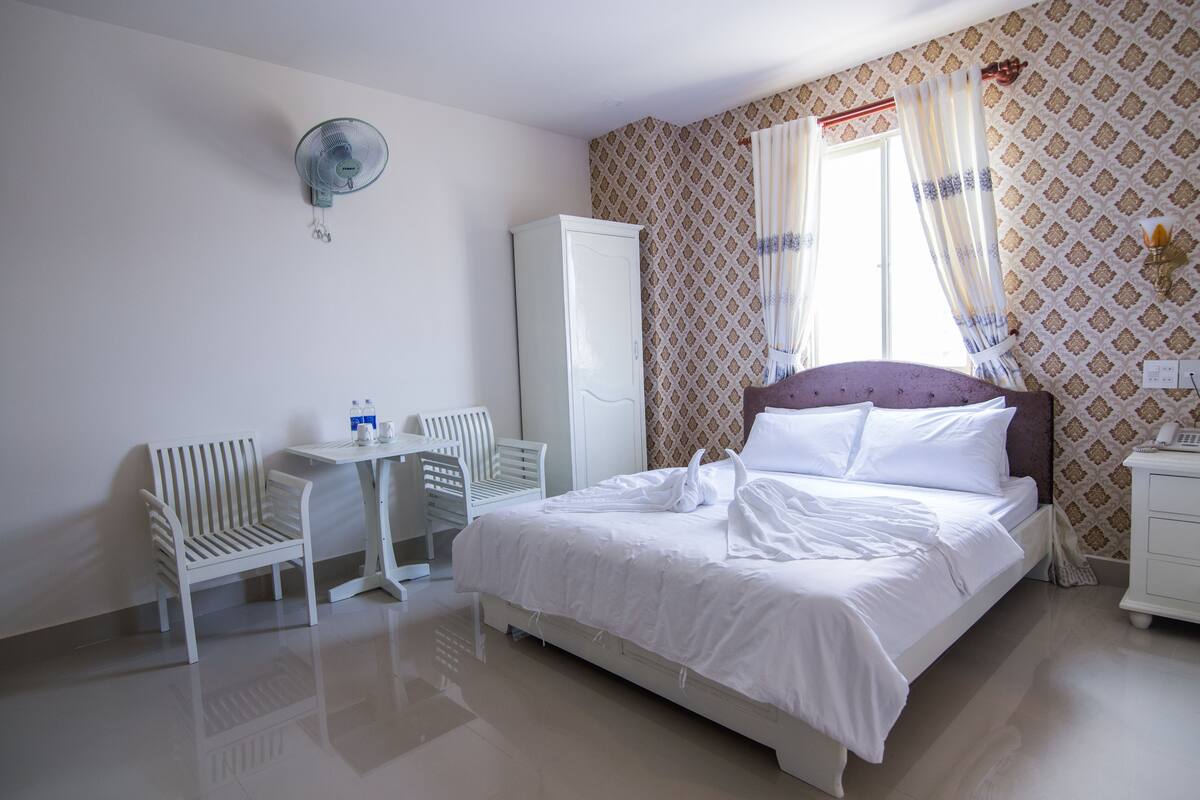 A neatly arranged hotel room with a bed, a small table with chairs, a wardrobe, and a fan.