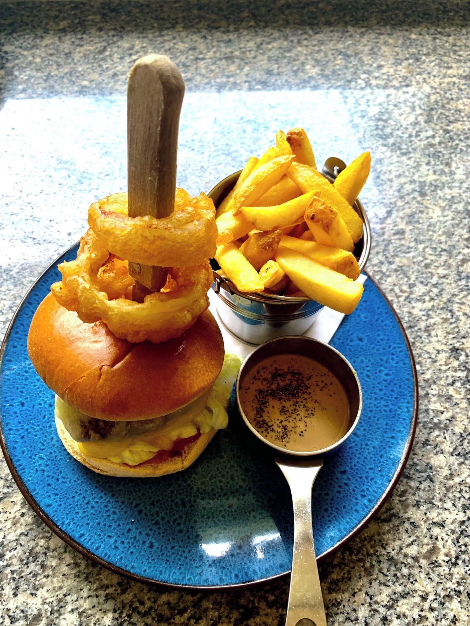A burger with onion rings, fries, and a dipping sauce on a blue plate.