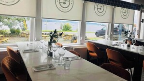 A restaurant with a view of the water, tables set with wine glasses and cutlery, and a logo on the wall. - The Grey Gull Hotel (Lochgilphead)
