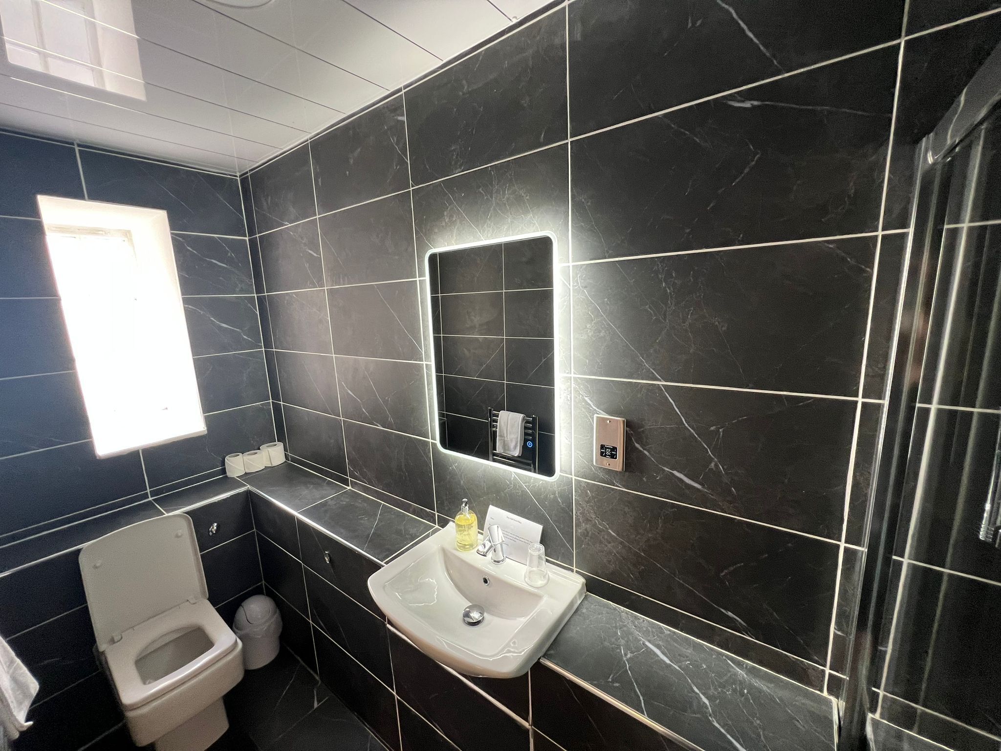 A modern bathroom with dark marble tiles, a white toilet, a sink with a mirror, and a small window.