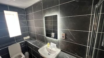 A modern bathroom with dark marble tiles, a white toilet, a sink with a mirror, and a small window.