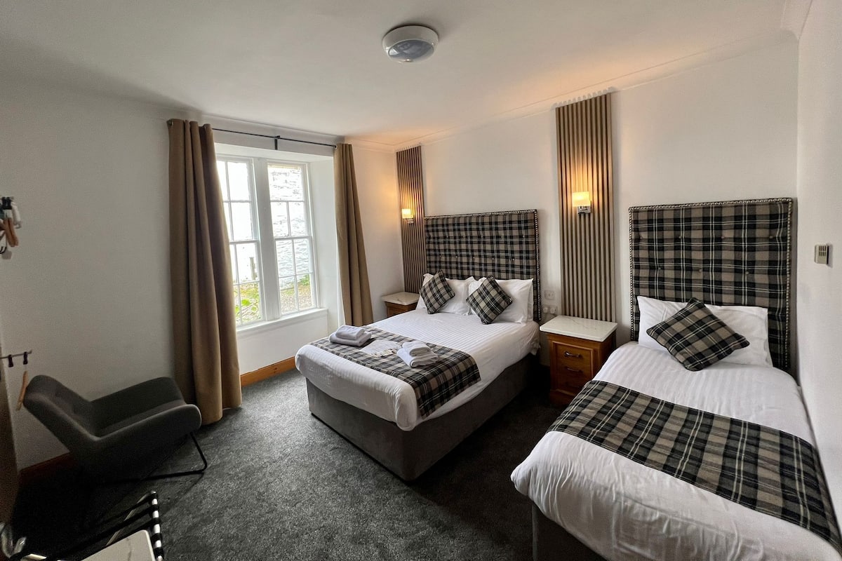 Une chambre d’hôtel avec deux lits, un bureau, une chaise, une fenêtre avec des rideaux et un luminaire fixé au mur.