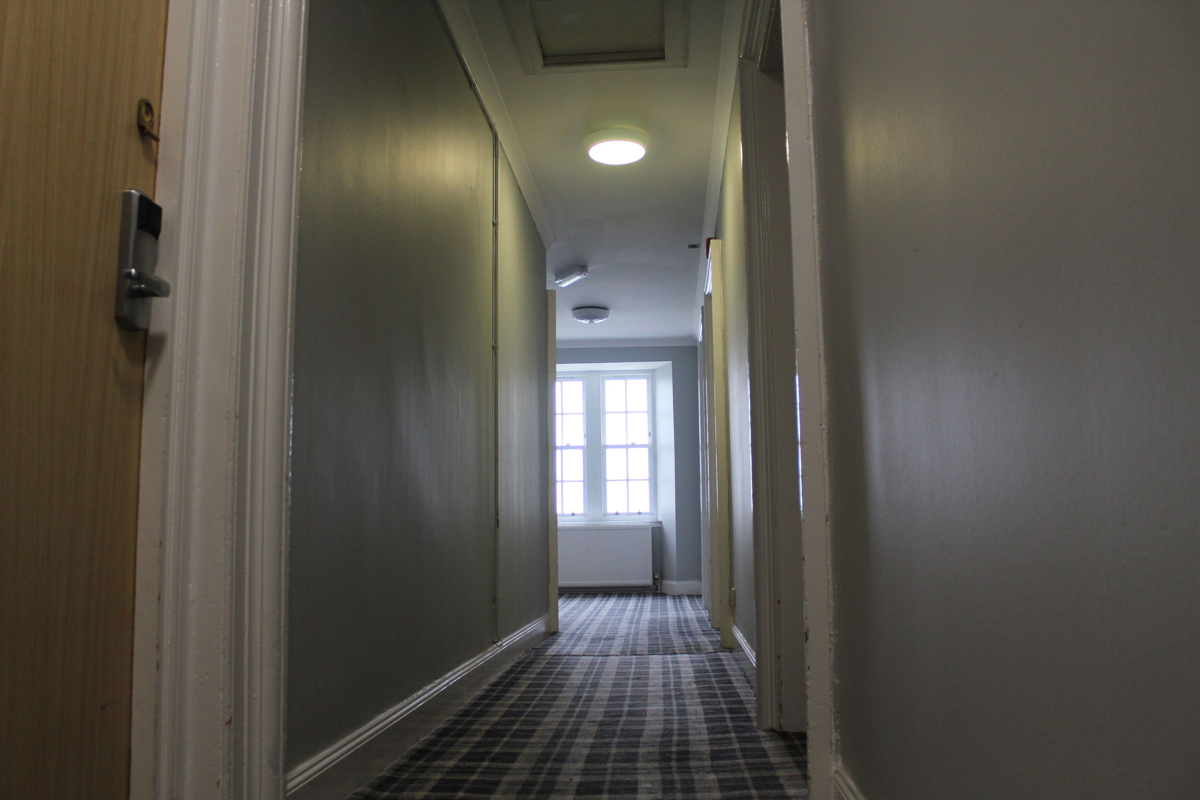 A narrow hallway with a checkered floor, a door on the left, and a window at the end.