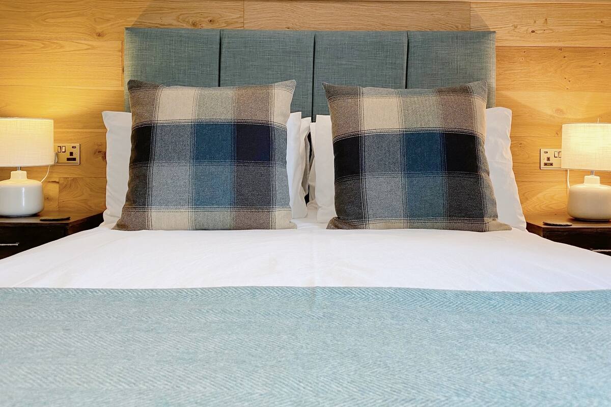 Una cama bien hecha con un almohadón de cuadros y una almohada blanca, flanqueada por dos mesitas de noche con lámparas.